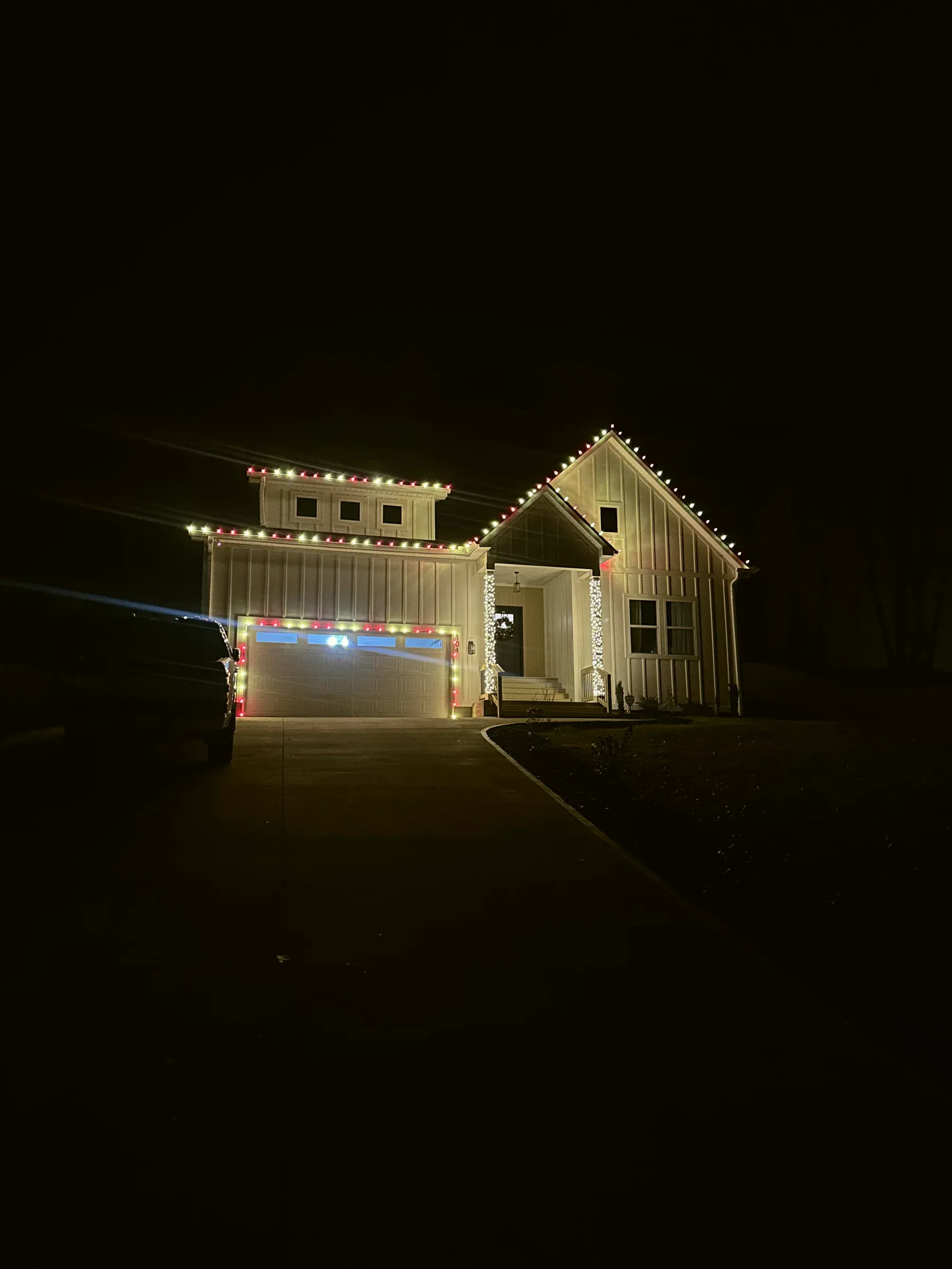 A board-and-batten house seen from the end of a long driveway at night, the roofline lit in red and white.