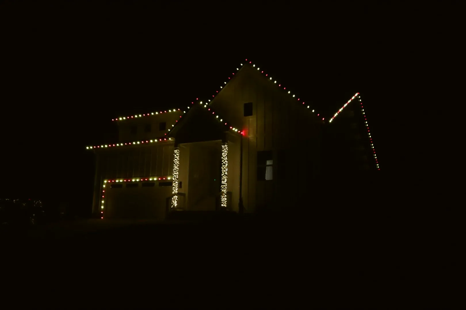 A board-and-batten house at night seen from across the lawn, its roofline, porch columns and garage outlined in red and white.