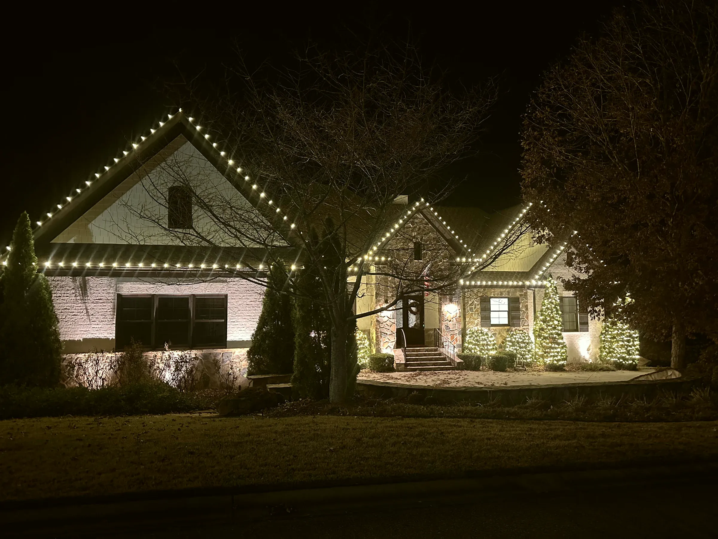 A stone and brick house at night with every gable outlined, the evergreens along the front wrapped and the trees out front lit.