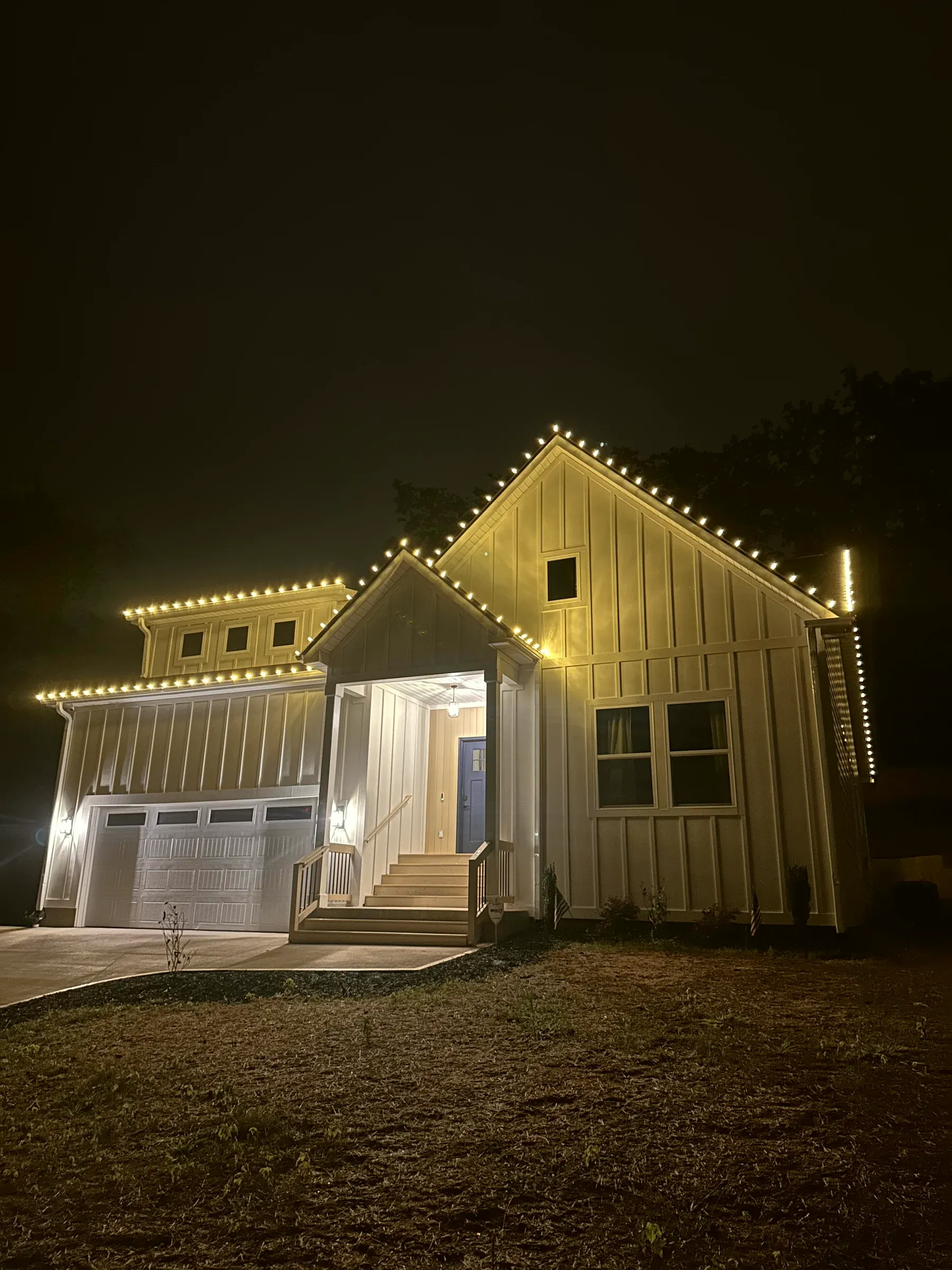 A board-and-batten house at night with its gables and porch roof outlined in warm white bulbs.